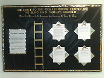 Bulletin board with black background and gold letters and along the border reads, Welcome to the Rehabilitation Center for the Blind and Visually Impaired Where our mission is the Independence and Employment of those we serve.