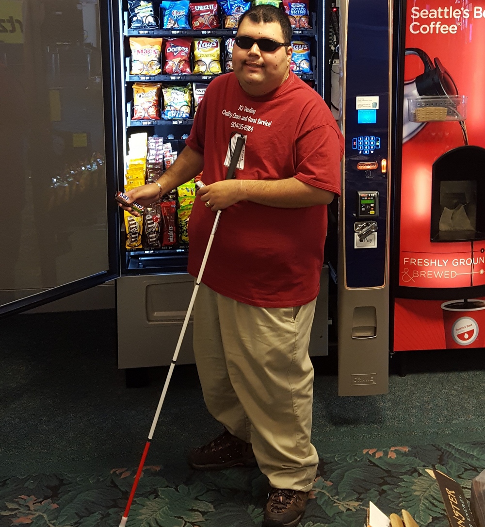 BBE vendor with a white cane and sunglasses standing next to a vending machine