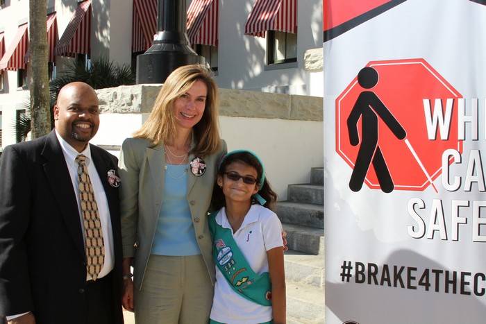 Three people smiling while standing next to a White Cane Safety Day sign
