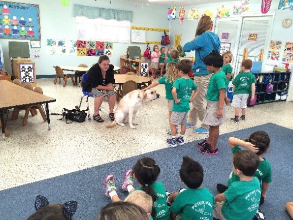 Children petting a service animal in a classroom while a woman speaks to them