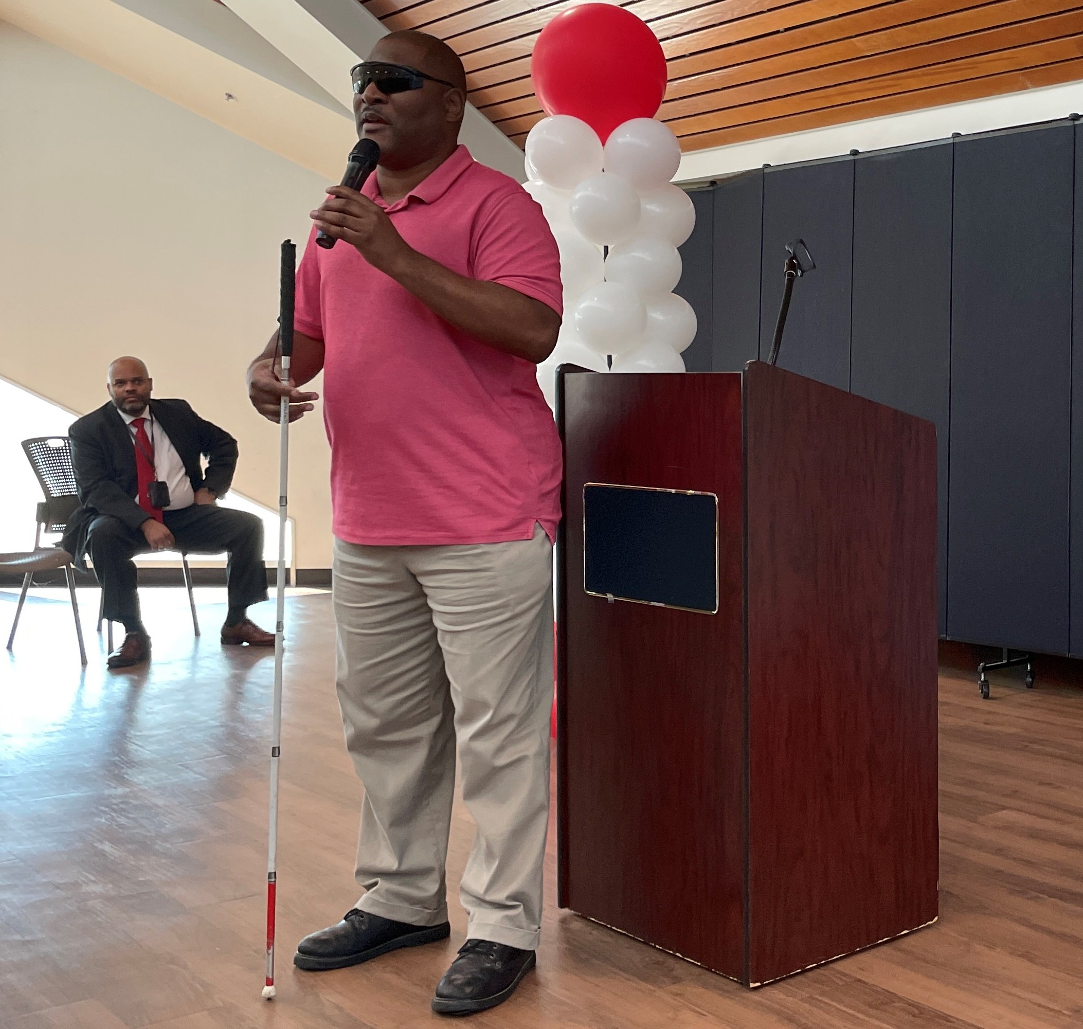 Guest speaker talks to the audience about using a white cane in front of white cane awareness banner and red and white balloons.