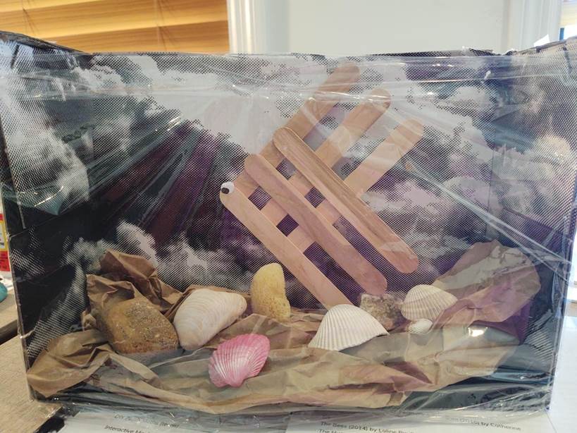 Shoebox aquarium, crumpled bag at bottom, rocks, shells, and craft stick angelfish, wrapped with clear plastic wrap.