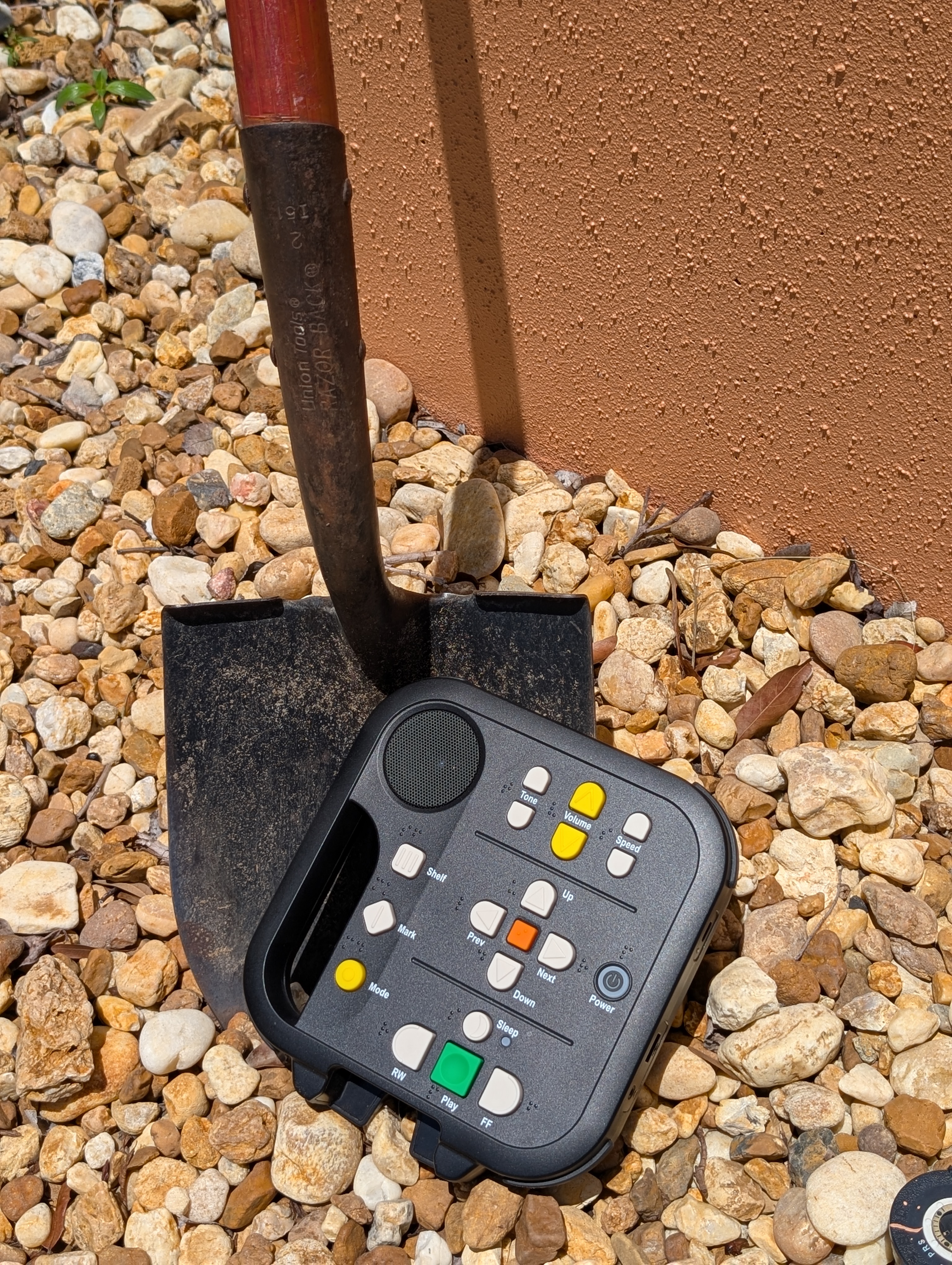 Rocks on the ground. A shovel on the rocks with a talking book player laying halfway off the shovel like it was just unearthed.