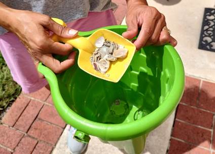 two hands holding a green beach bucket with a yellow shovel with a bunch of small seashells, covered in sand.